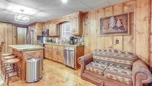 A kitchen with wooden cabinets, a stainless steel dishwasher, a bar stool area, and a patterned sofa. - Wooded Blue Ridge Cabin: 2 Decks, Fire Pit! (Cherry Log)