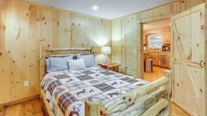 A wooden-paneled bedroom with a bed, a nightstand, and a view into a kitchen area. - Wooded Blue Ridge Cabin: 2 Decks, Fire Pit! (Cherry Log)