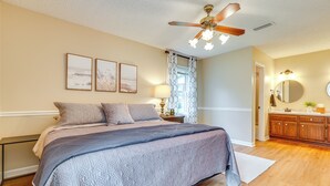 A bedroom with a bed, a ceiling fan, a nightstand with a lamp, a window with curtains, and a bathroom with a mirror and sink. - Decatur Vacation Rental w/ Patio! (Decatur)