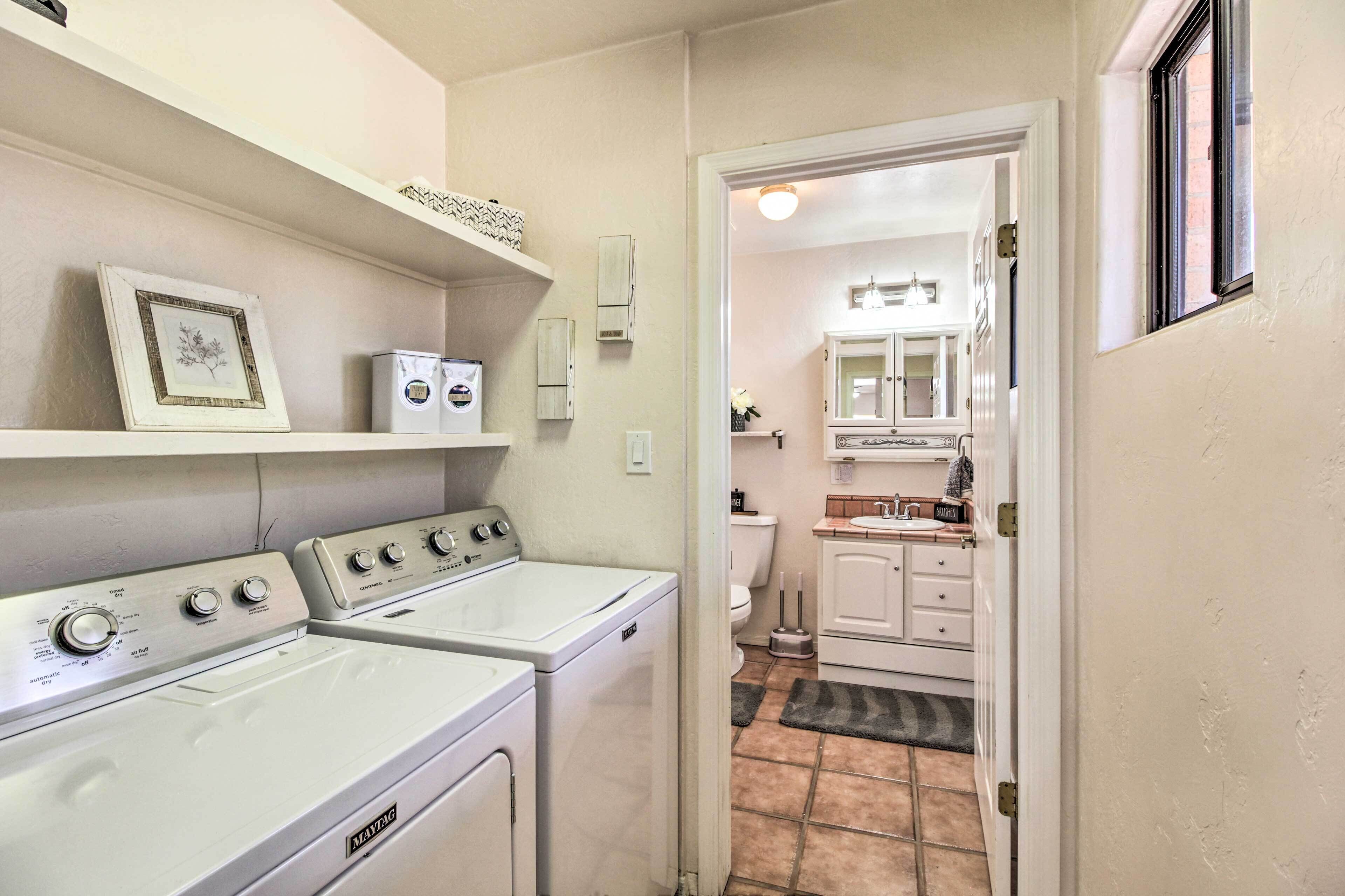 A laundry room with two front-loading washing machines and a dryer, a small shelf with decorative items, and a view into the adjacent bathroom with a toilet and sink.