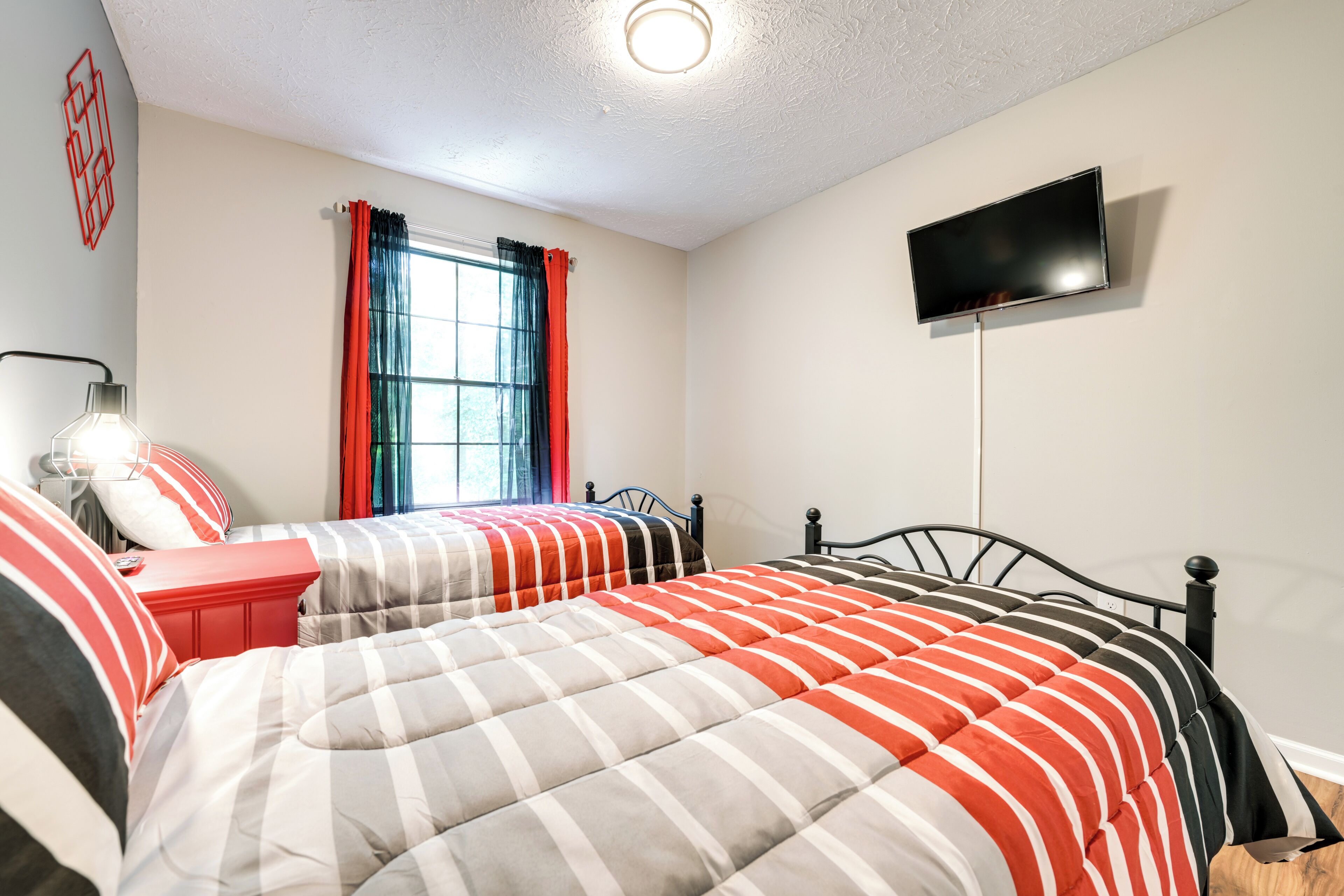 A bedroom with two beds, a TV, a window with curtains, and a bedside table with a lamp.