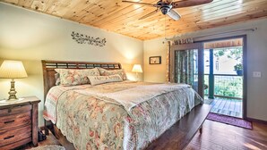 A bedroom with a wooden ceiling, a bed with floral bedding, a nightstand with a lamp, and a view of the outdoors through a door.