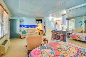 A room with a bed, a chair, a desk, a television, and a kitchen area.