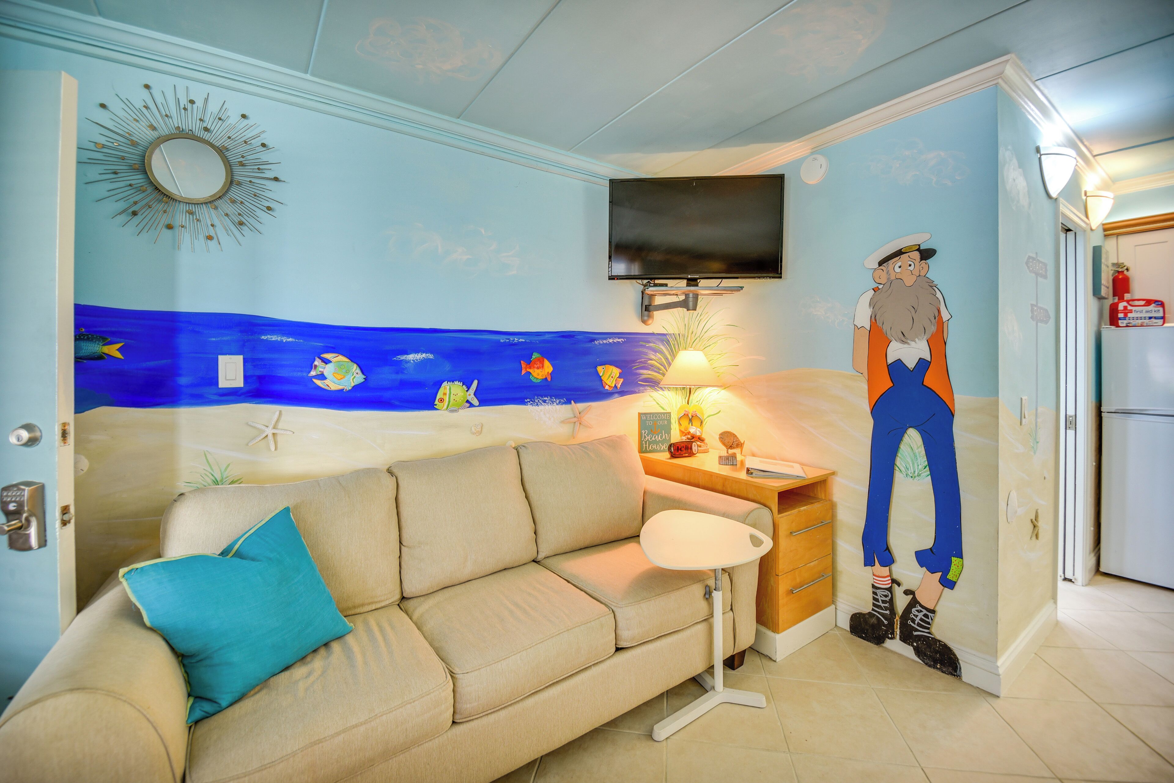 A living room with a sofa, a TV mounted on the wall, a lamp, and a mural of a beach scene.