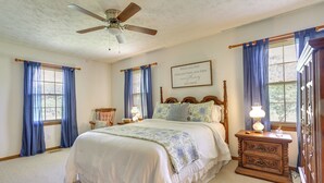 A bedroom with a large bed, a wooden nightstand, a ceiling fan, and a window with blue curtains. - Cozy Georgia Getaway w/ Deck, Grill & Fire Pit! (Cartersville)