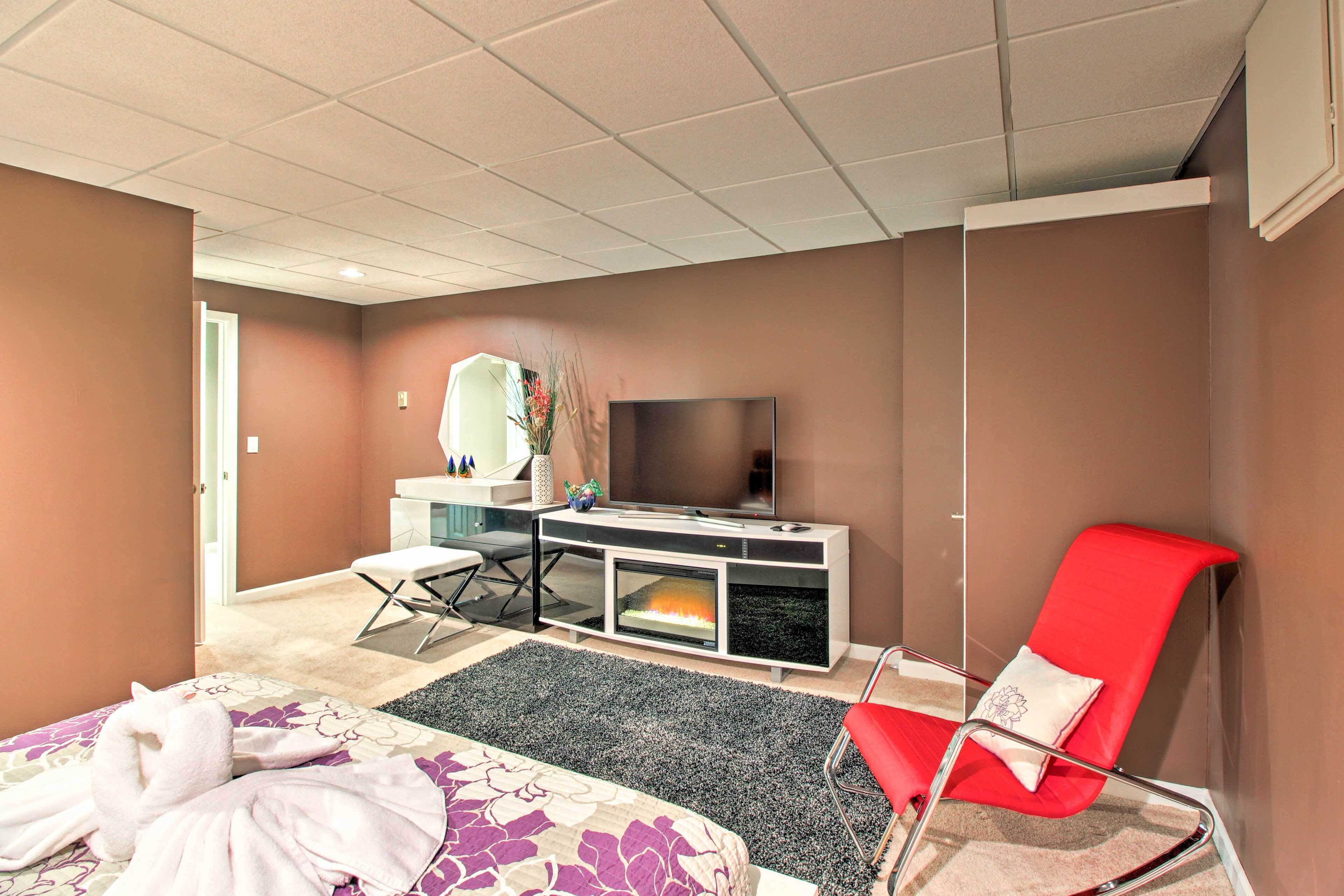 A hotel room with a bed, a TV, a red chair, a desk with a flat-screen TV, and a fireplace.