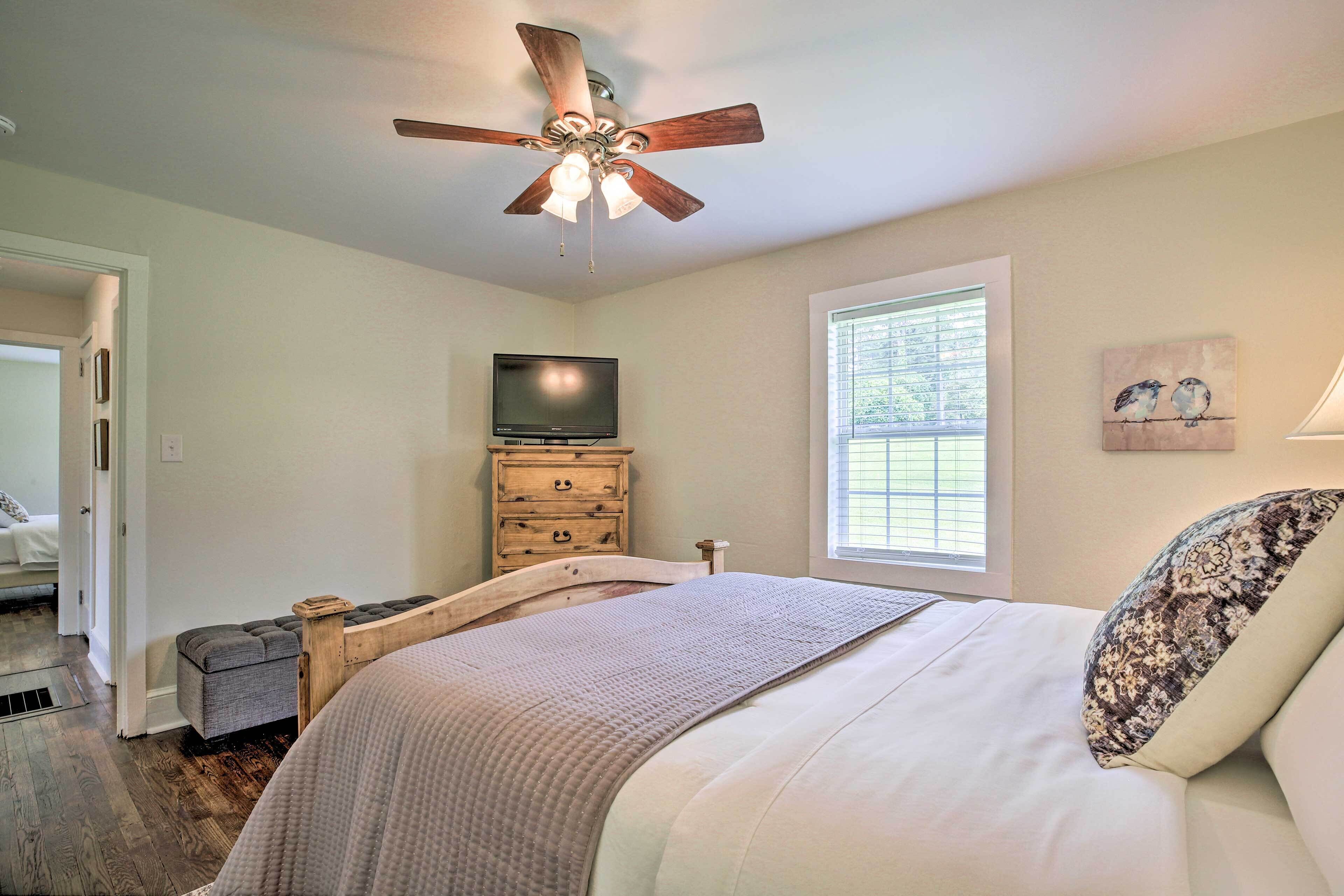 A bedroom with a bed, a ceiling fan, a television, a window, and a painting on the wall.