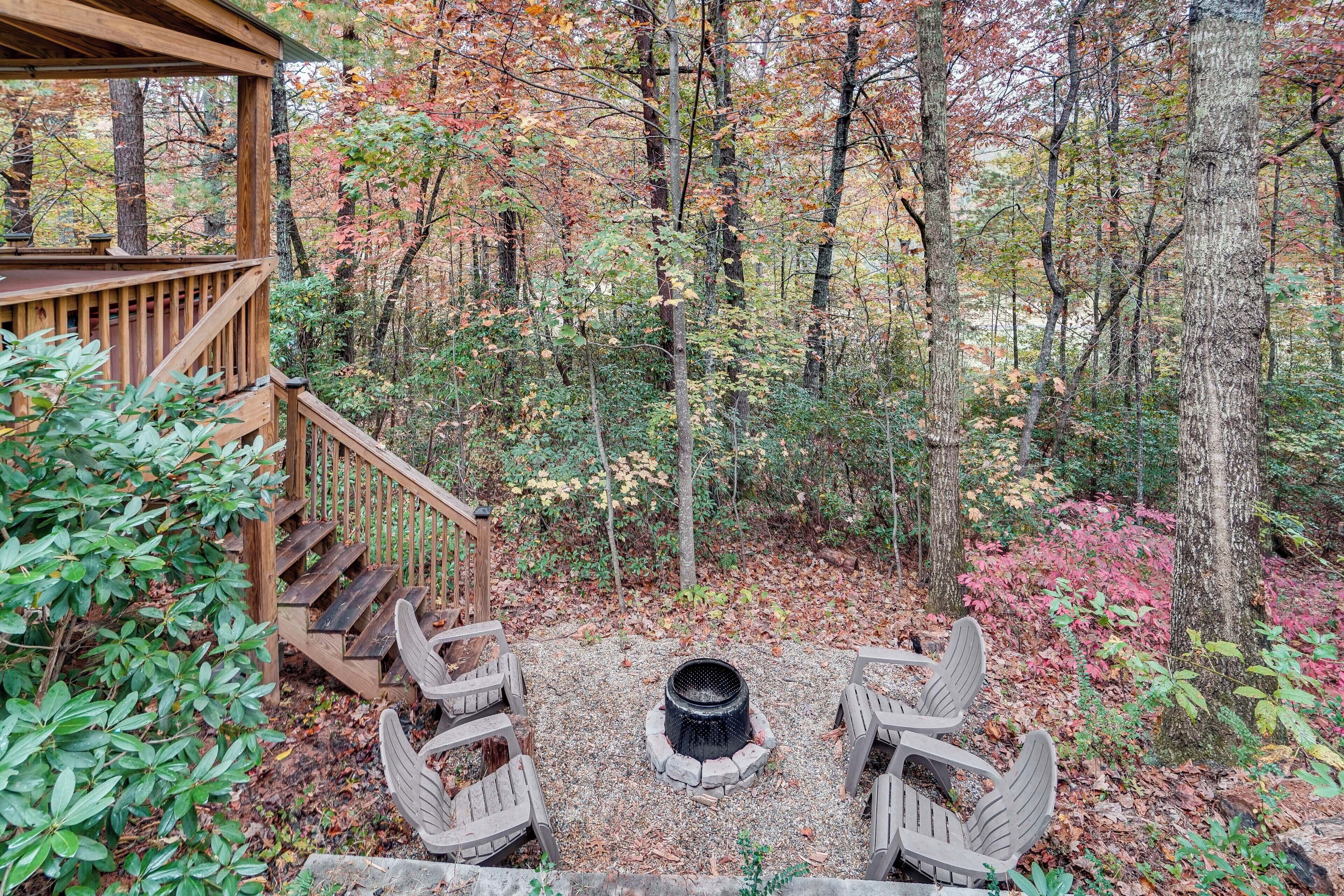 A wooden deck with a fire pit and Adirondack chairs in a forested area.