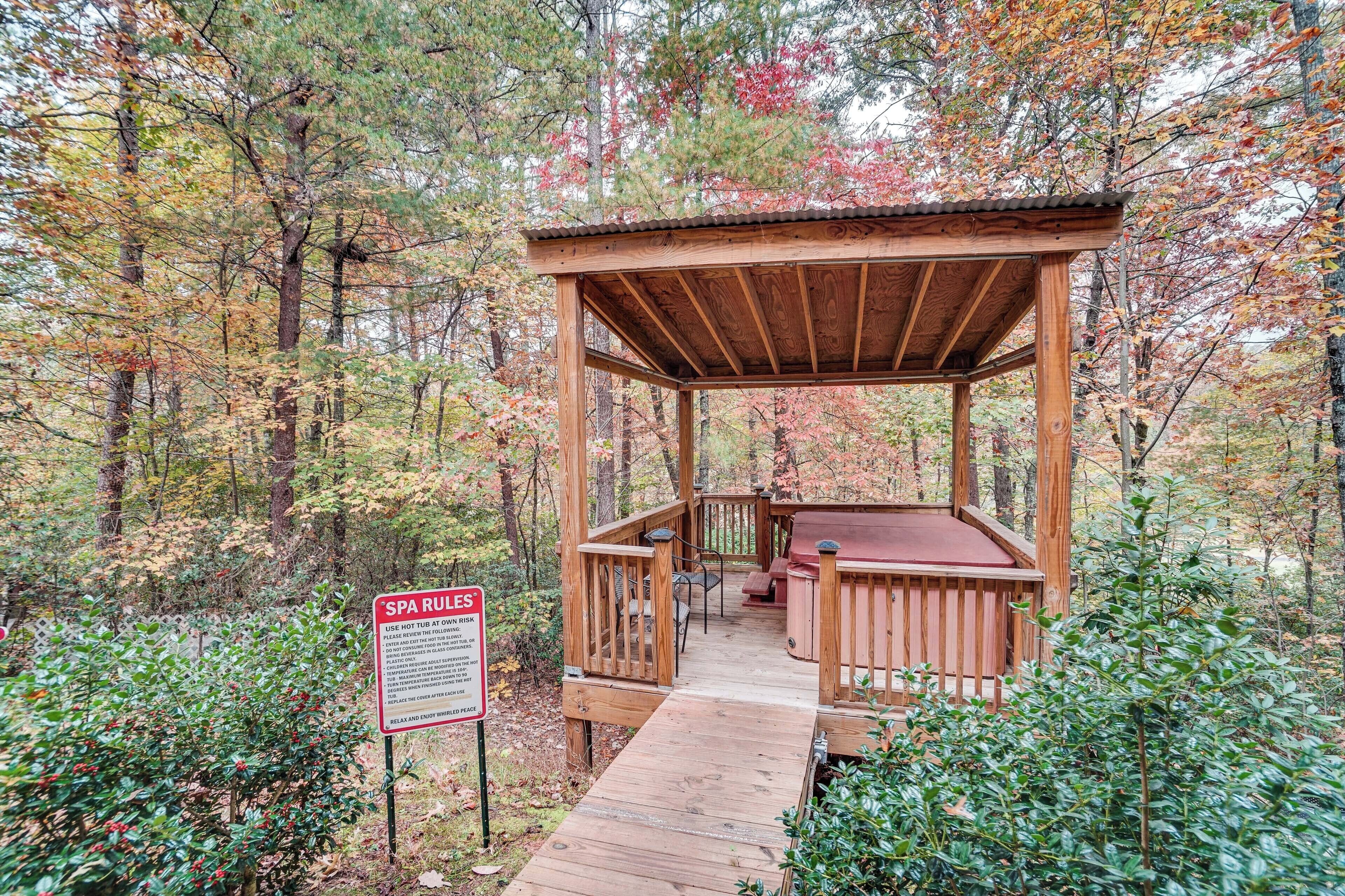 A wooden gazebo with a bench and a hot tub in a forested area.