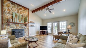 A spacious living room with a stone fireplace, wooden beams, a ceiling fan, and a large window. - Lake Sinclair Retreat w/ Big Water Views & Dock! (Milledgeville)