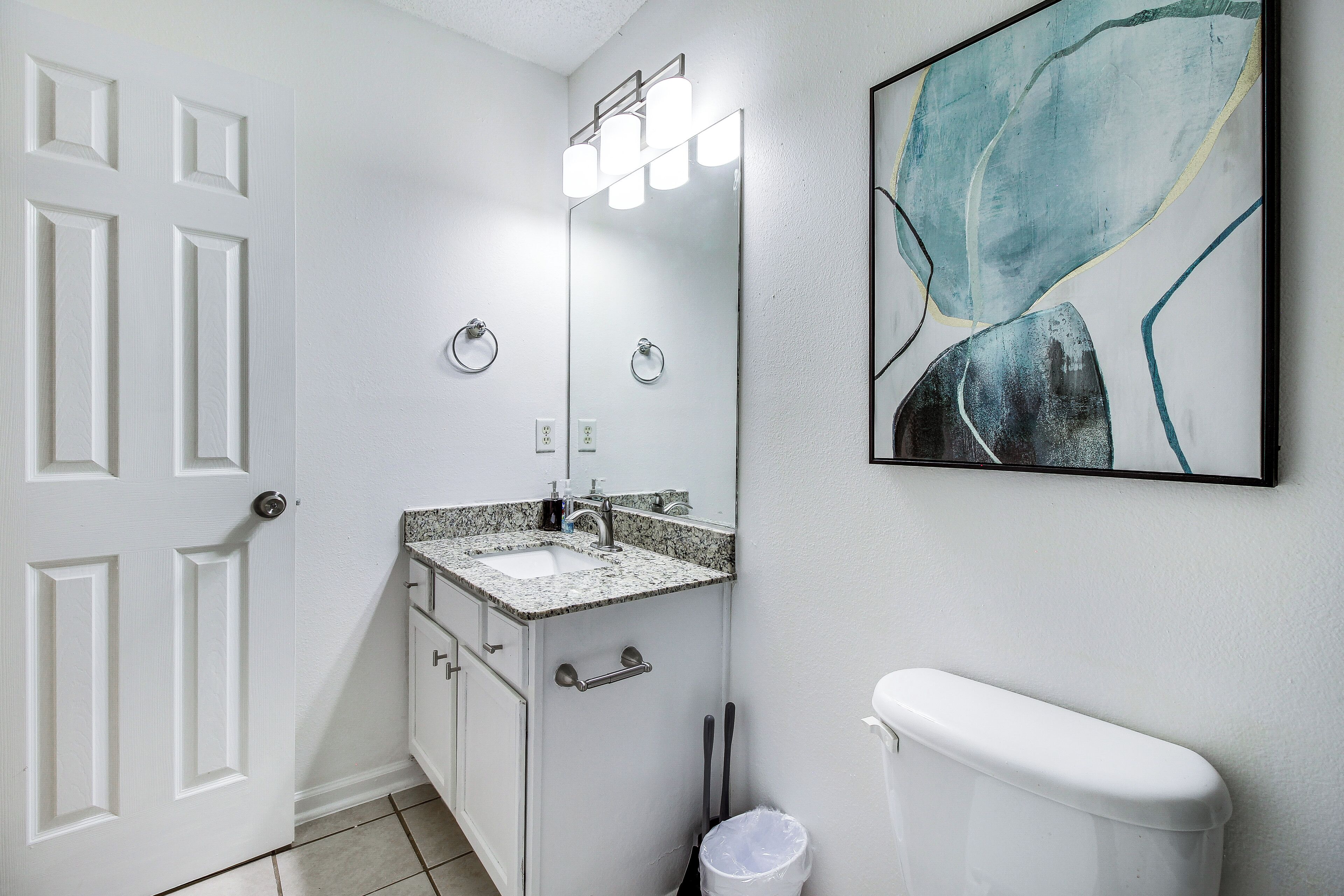 A bathroom with a white door, a sink with a mirror, a toilet, and a trash can.