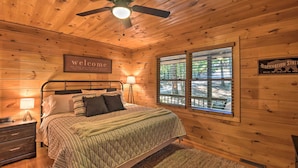 Ein Schlafzimmer einer Holzhütte mit Bett, Nachttisch, Lampe, Fenster mit Aussicht und einem 'Welcome'-Schild.