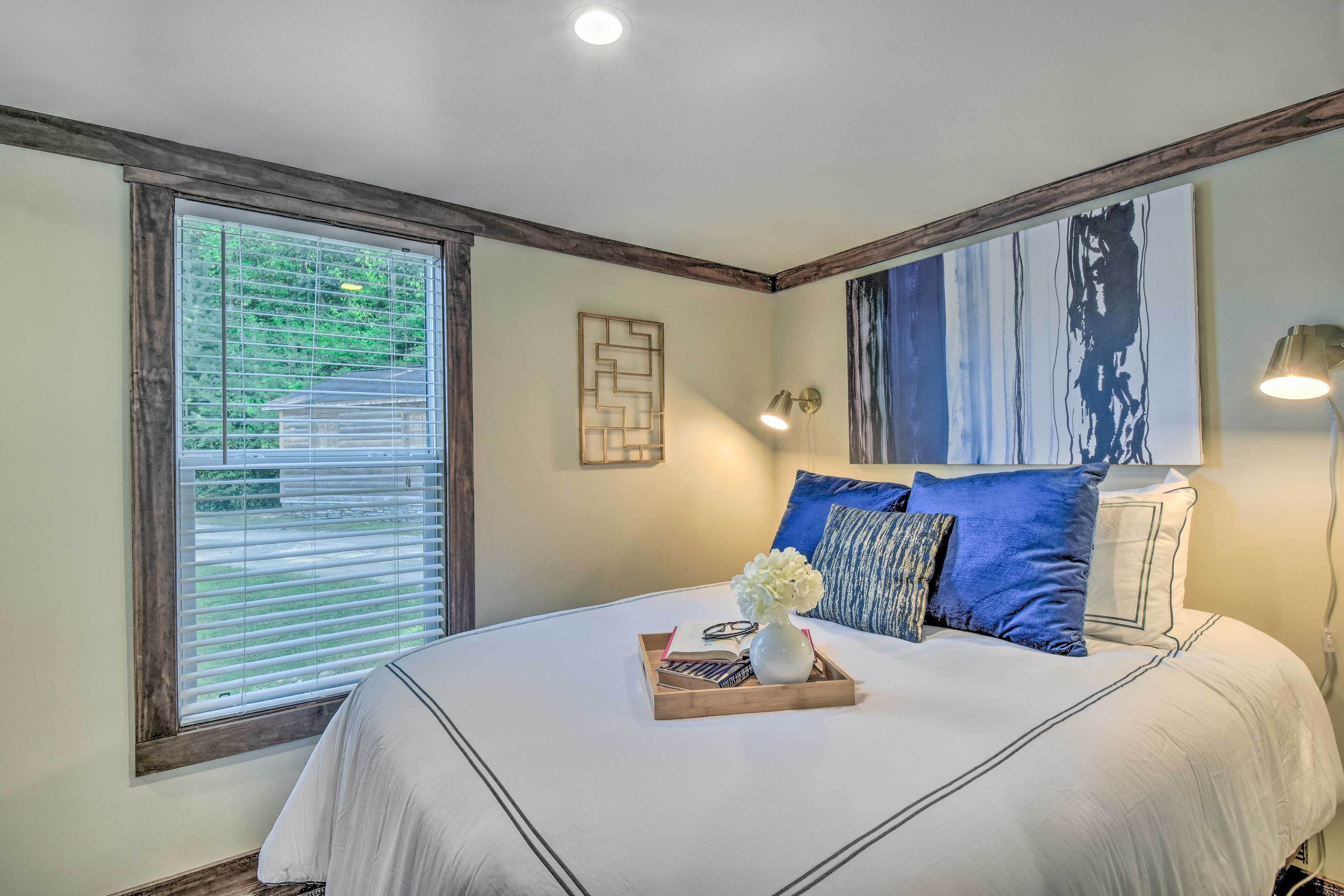 A bedroom with a large bed, a window with blinds, a wall art piece, and a bedside lamp.