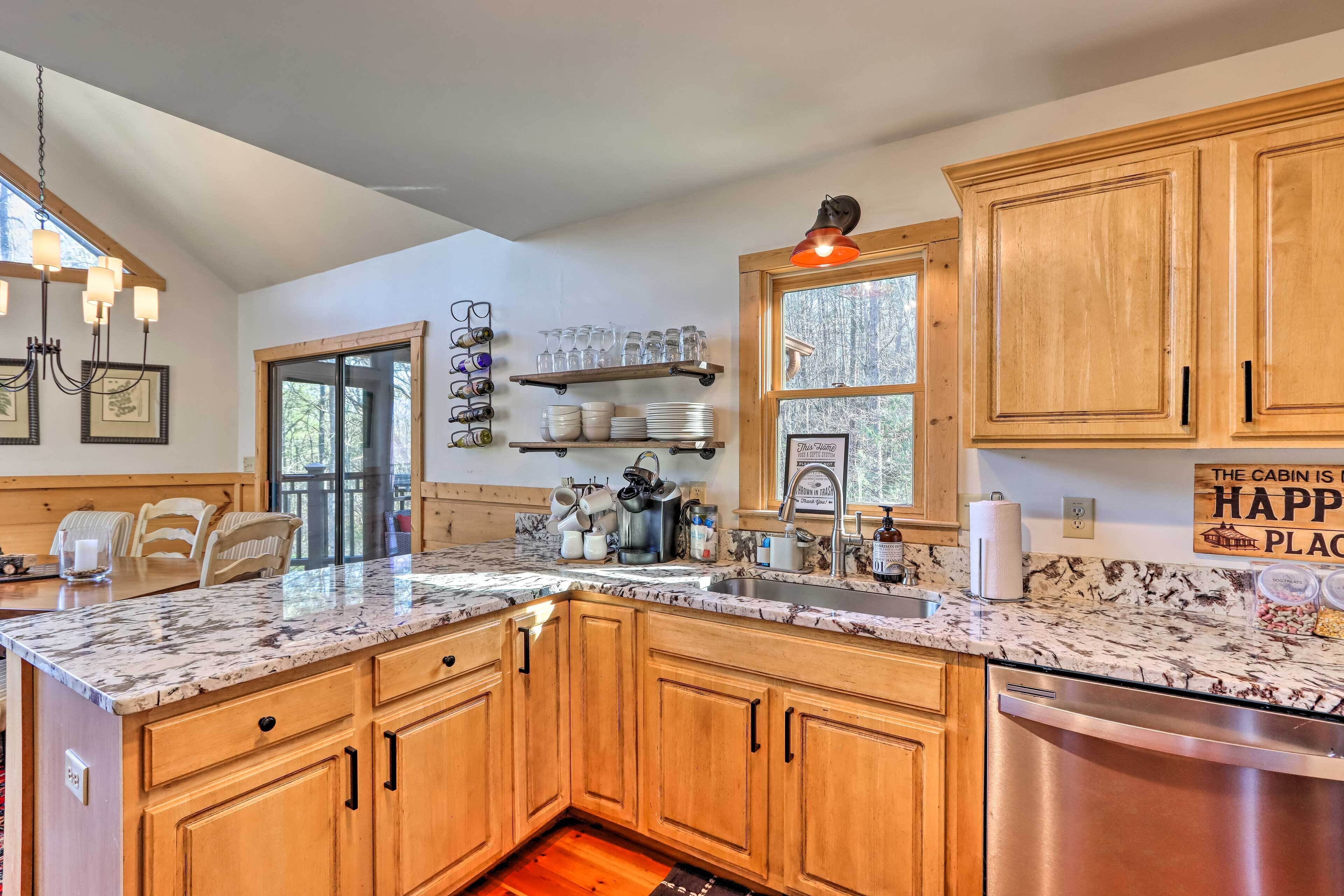 A kitchen with wooden cabinets, a granite countertop, and a stainless steel dishwasher. There are shelves with glassware and plates, and a window with a view of trees.