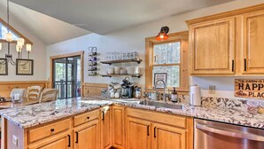 A kitchen with wooden cabinets, a granite countertop, and a stainless steel dishwasher. There are shelves with glassware and plates, and a window with a view of trees. - Spacious Ellijay Resort Home w/ Hot Tub! (Ellijay)