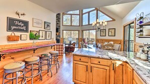 A spacious kitchen with a large island, wooden cabinets, and a stone accent wall. There are multiple stools and a bench along the counter, and a dining area with a table and chairs visible through the large windows. - Spacious Ellijay Resort Home w/ Hot Tub! (Ellijay)