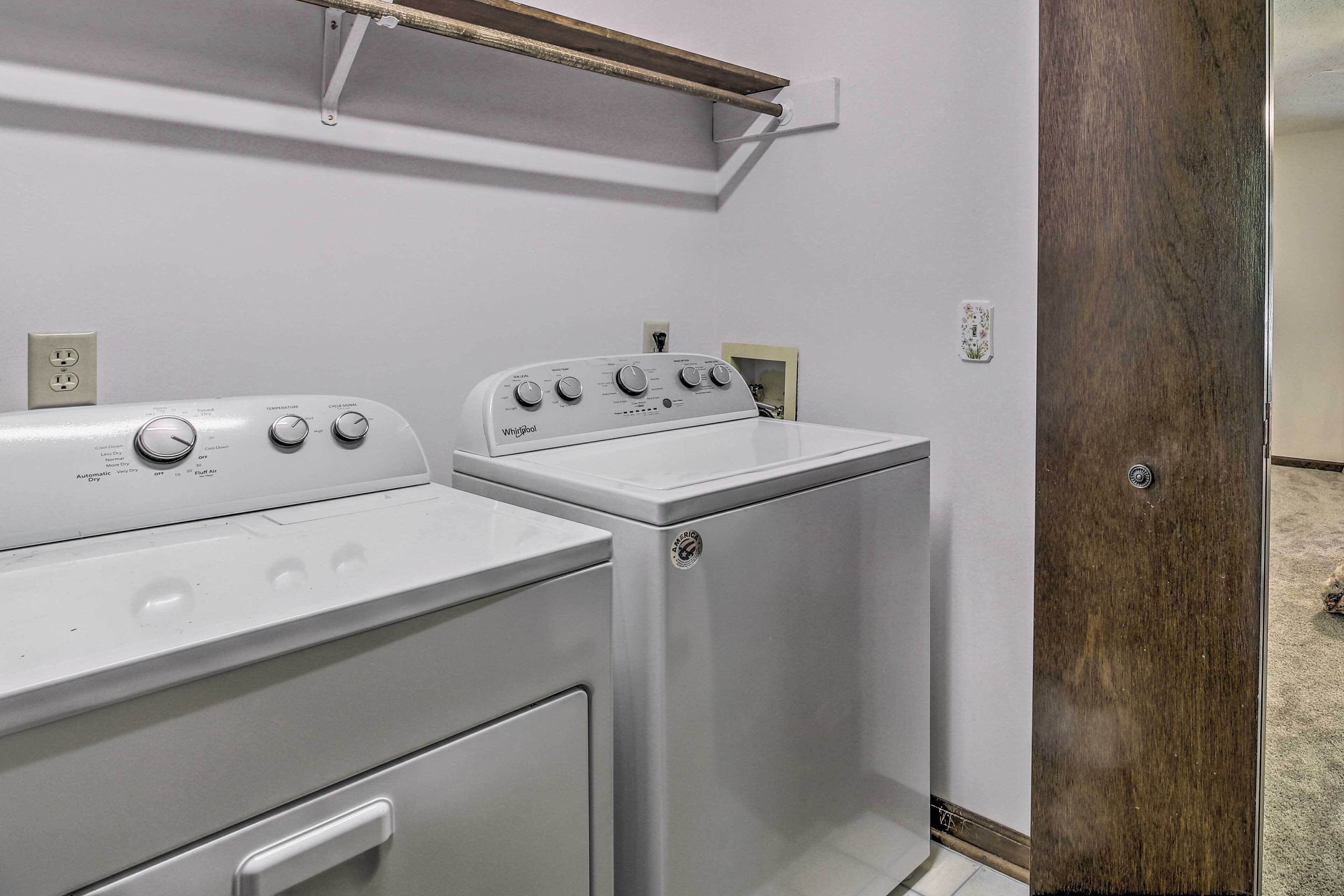 A laundry room with a white washer and dryer, a wooden shelf above the dryer, and a door to the right.