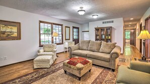A spacious living room with a large sofa, a smaller armchair, and a matching ottoman. There are two windows with curtains, a wooden cabinet, and a lamp on a side table.