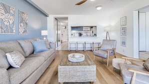 A living room with a sofa, coffee table, and chairs, a kitchen in the background, and a ceiling fan. - June Specials! Beautiful Stylish Condo W/ Amazing Ocean Views! (Kihei)