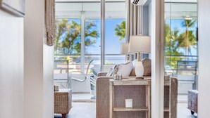 A modern living room with a sofa, a lamp, and a view of palm trees through large windows. - June Specials! Beautiful Stylish Condo W/ Amazing Ocean Views! (Kihei)