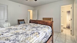 Une chambre à coucher avec un lit, un ventilateur de plafond, une tête de lit en bois, une table de chevet et une salle de bain visible à travers une porte ouverte.