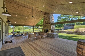 An outdoor patio with wooden flooring, wicker furniture, a stone fireplace, and a view of the surrounding landscape.