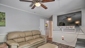 A living room with a beige sofa, an ottoman, a ceiling fan, a flat-screen TV, and a wooden TV stand. - Spacious Retreat ~ 1 Mile to Augusta National! (Augusta)