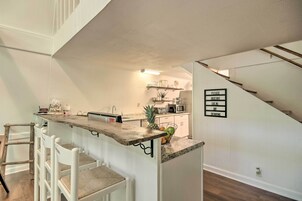 A kitchen with a wooden bar counter, white chairs, a microwave, and a sign instructing to observe social distancing.