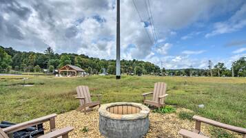 A circular fire pit surrounded by wooden Adirondack chairs, set on a gravel area with a grassy field and trees in the background.