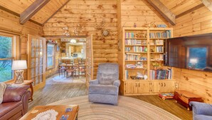 A cozy log cabin interior with a fireplace, bookshelves, a comfortable armchair, and a dining area.