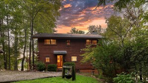 A rustic log cabin with a wooden porch, surrounded by dense trees and a clear sky.
