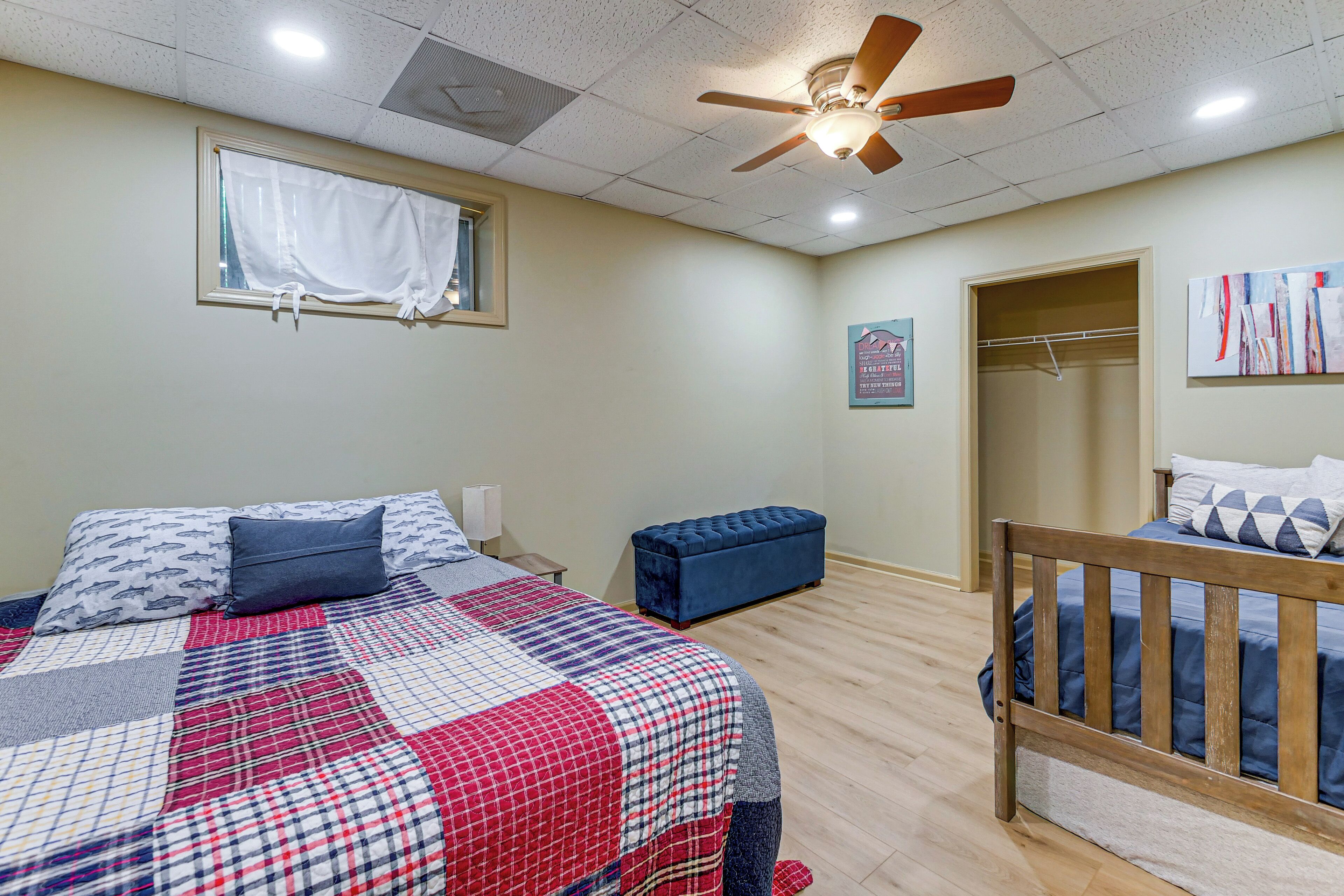 A bedroom with a bed, a wooden crib, a blue bench, a ceiling fan, a window with curtains, and a framed picture on the wall.