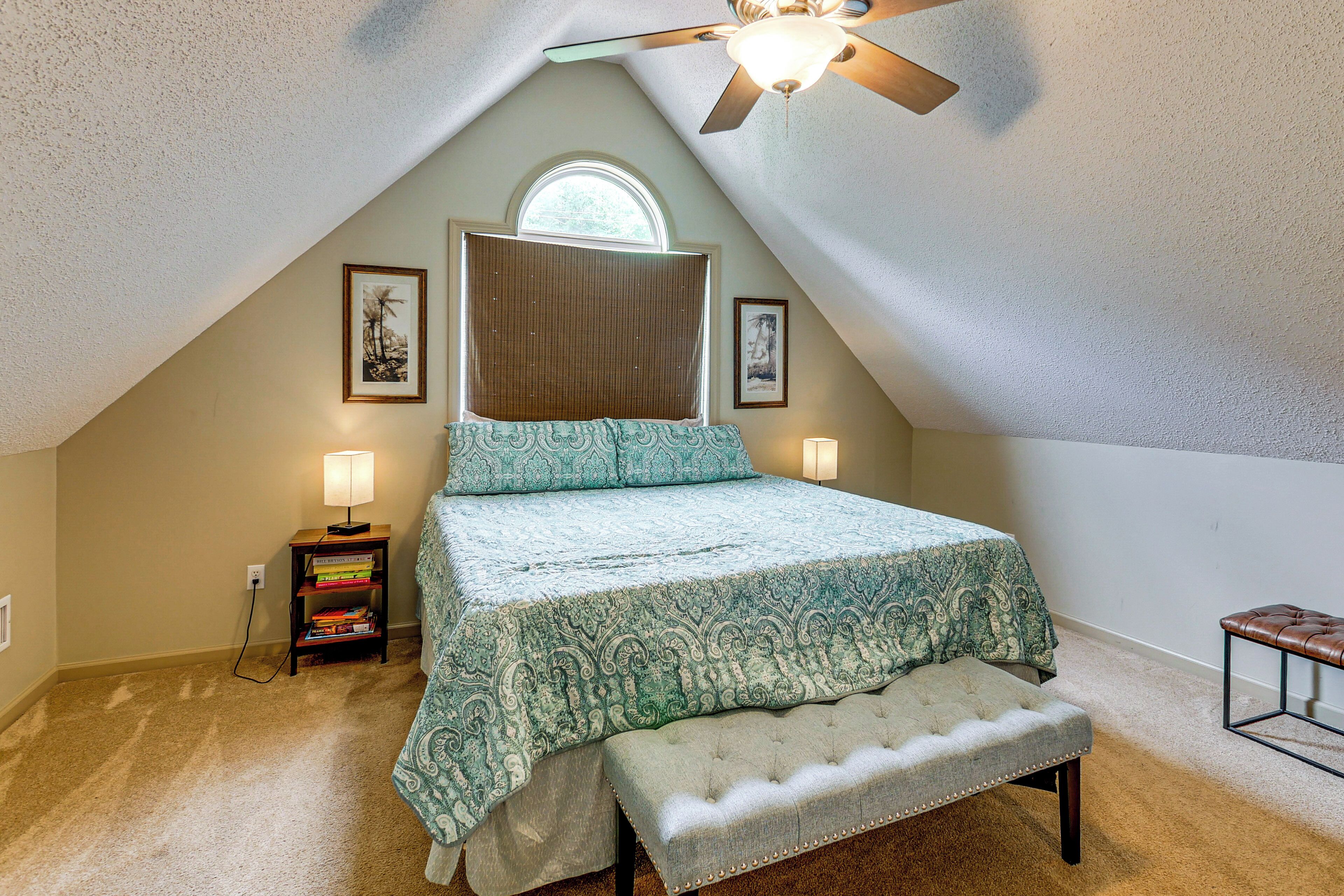 An attic bedroom with a sloped ceiling, a bed with a patterned bedspread, a bench, a nightstand with books, and a ceiling fan.
