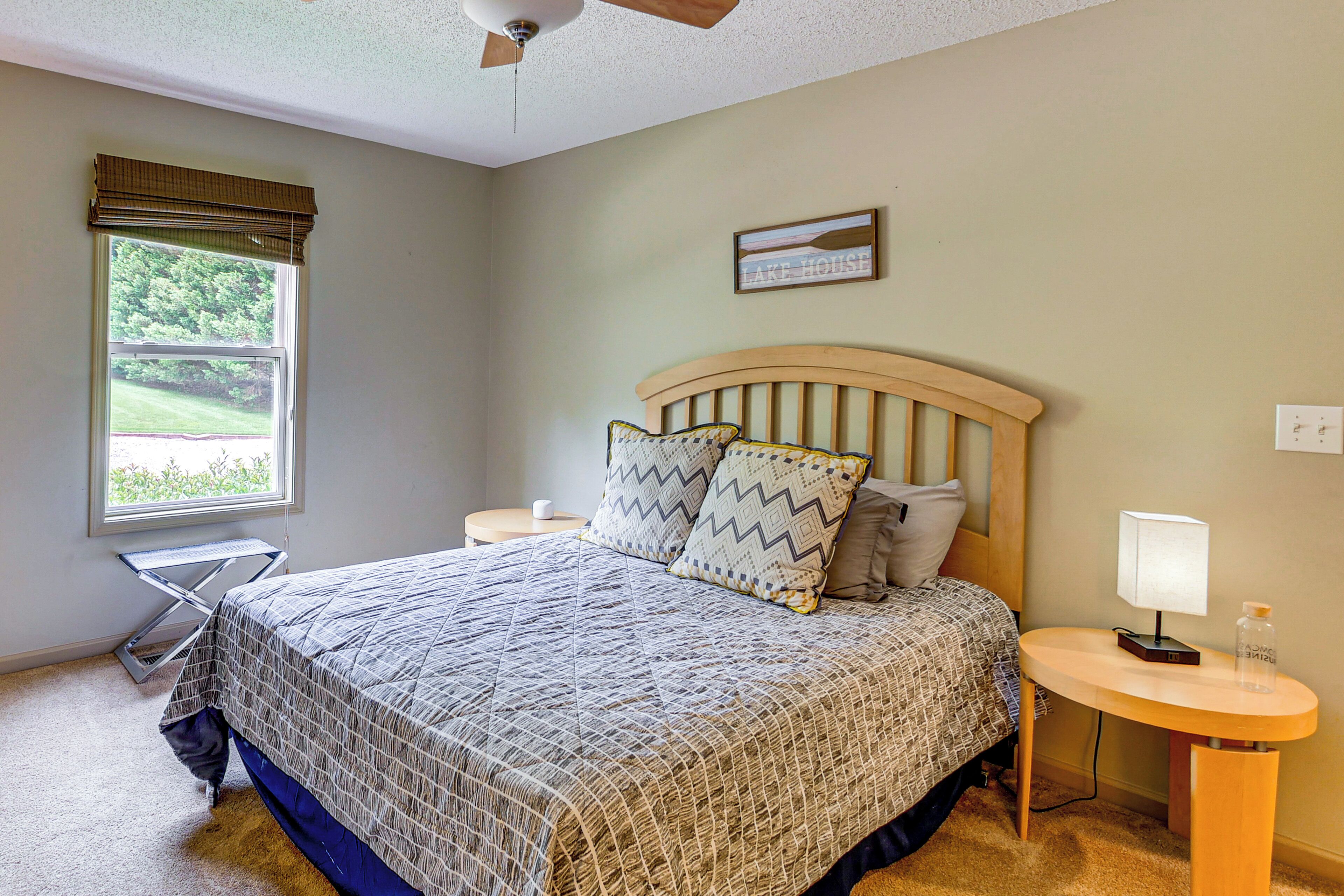 A bedroom with a bed, a bedside table with a lamp, a window with blinds, and a ceiling fan.