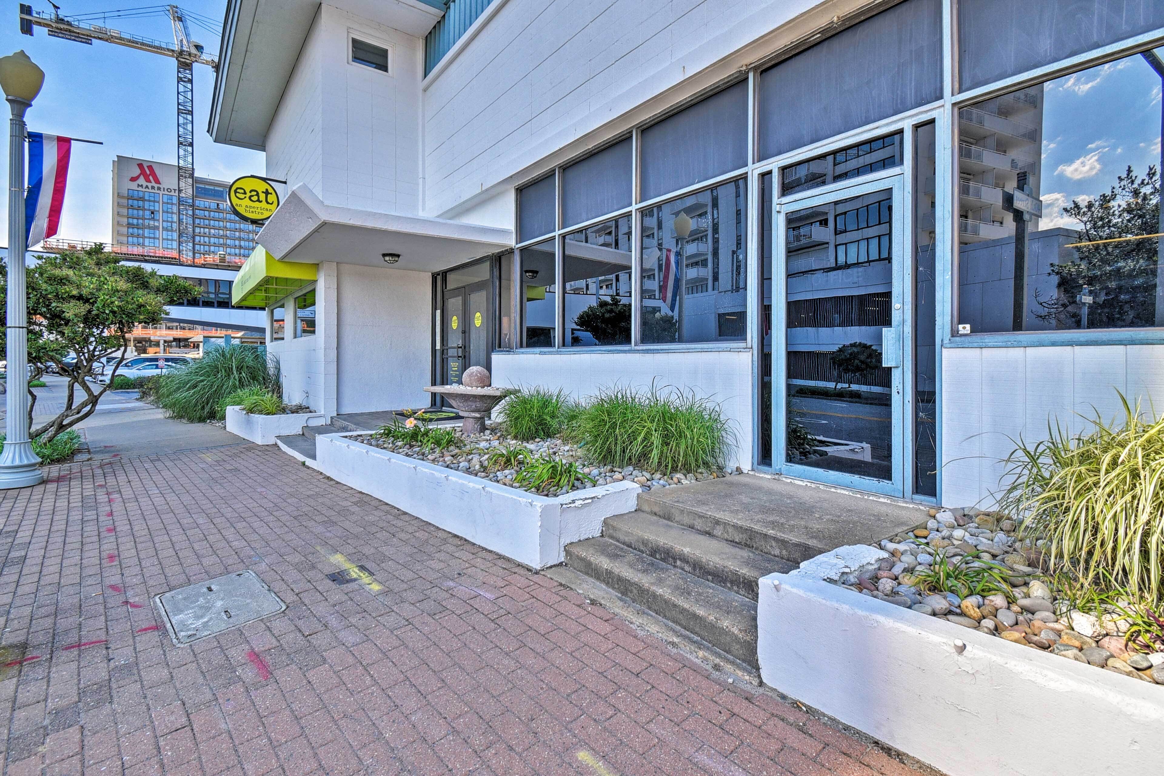 A modern building with a large glass entrance, a sign indicating a restaurant, and a landscaped area with plants and steps leading to the entrance.