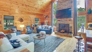 A cozy log cabin interior with a stone fireplace, wooden ceiling, and a large TV mounted on the wall. - Upscale Coosawattee Cabin w/ Hot Tub & Fire Pit! (Ellijay)