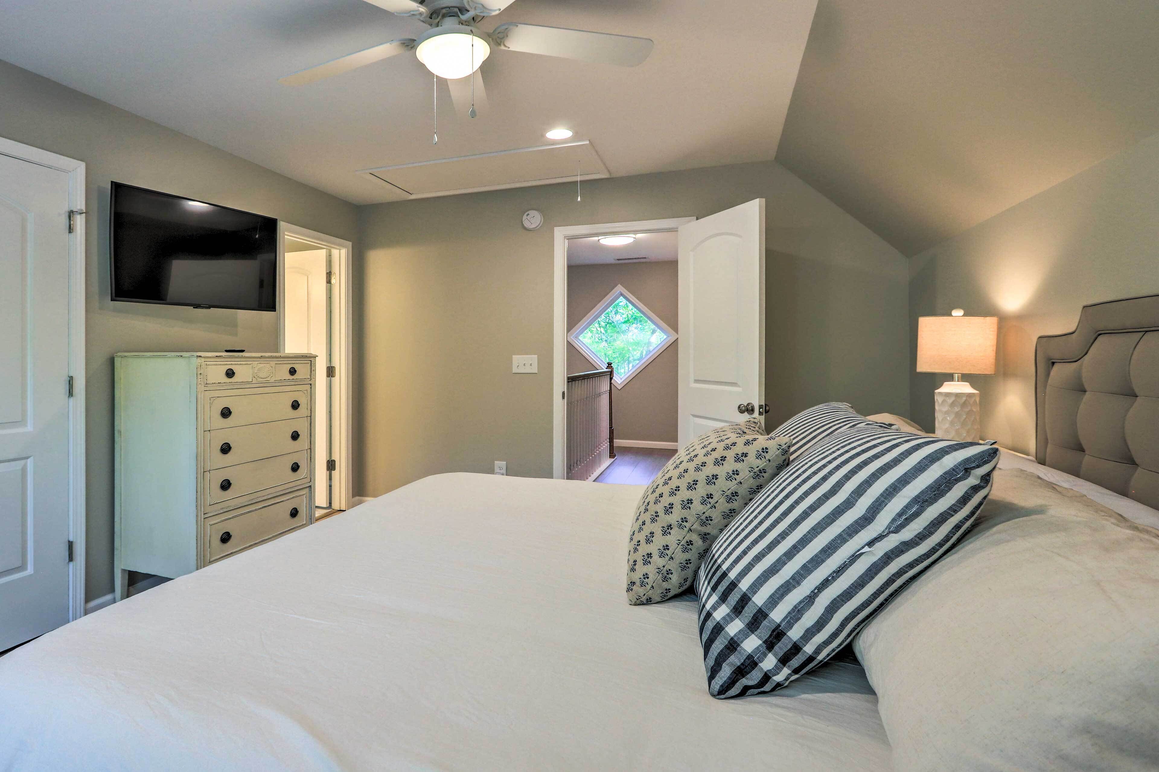 Une chambre avec un lit, un ventilateur de plafond, un téléviseur fixé au mur, une commode et une fenêtre donnant sur un paysage verdoyant.