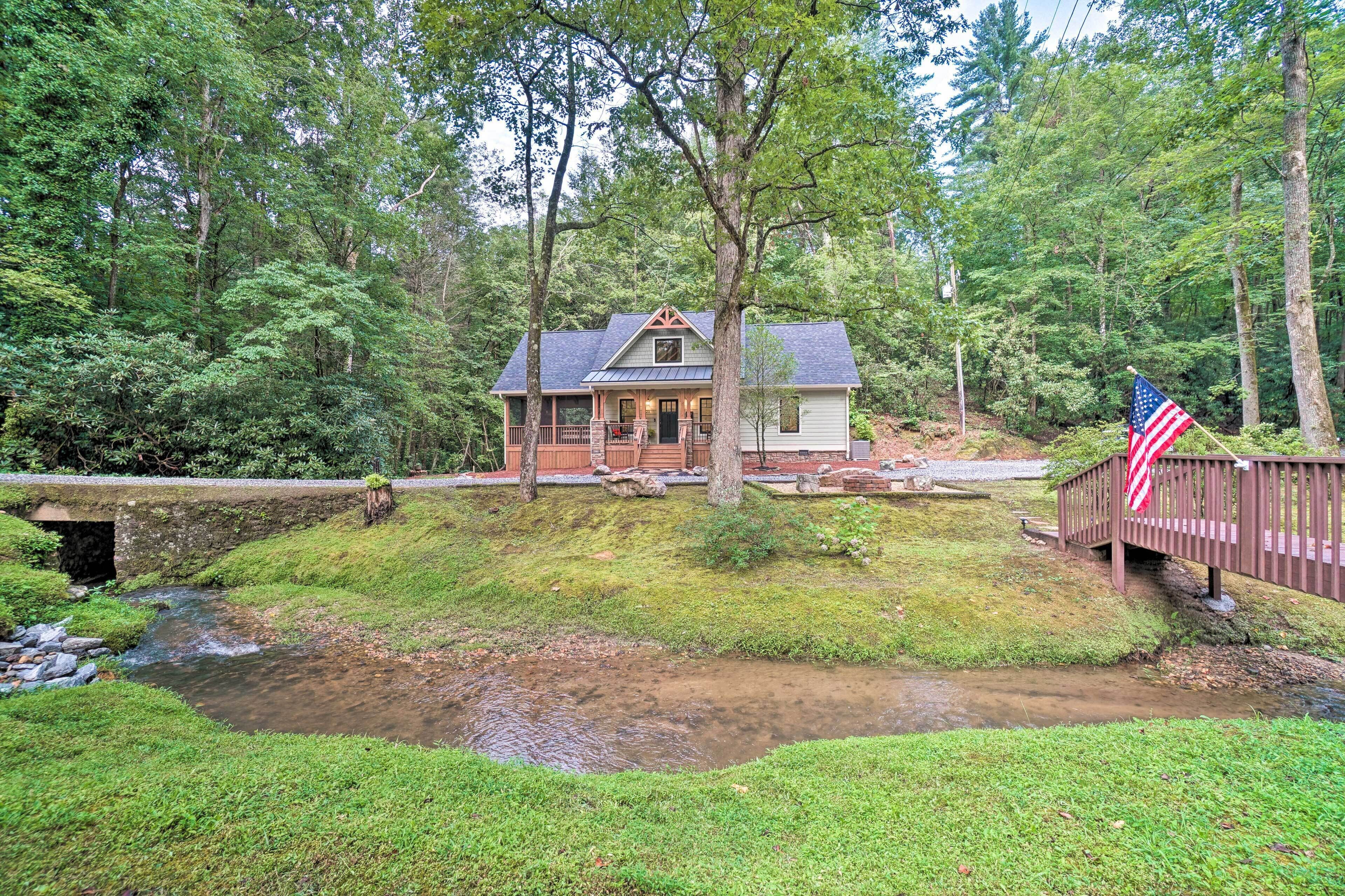 A cabin with a porch, surrounded by a forest and a small stream.