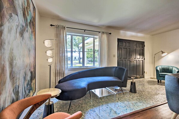 A modern living room with a curved sofa, a round coffee table, and a floor lamp. A large window with curtains allows natural light to enter. - Ocean Springs Home - Walk to Beach & Downtown! (Ocean Springs)