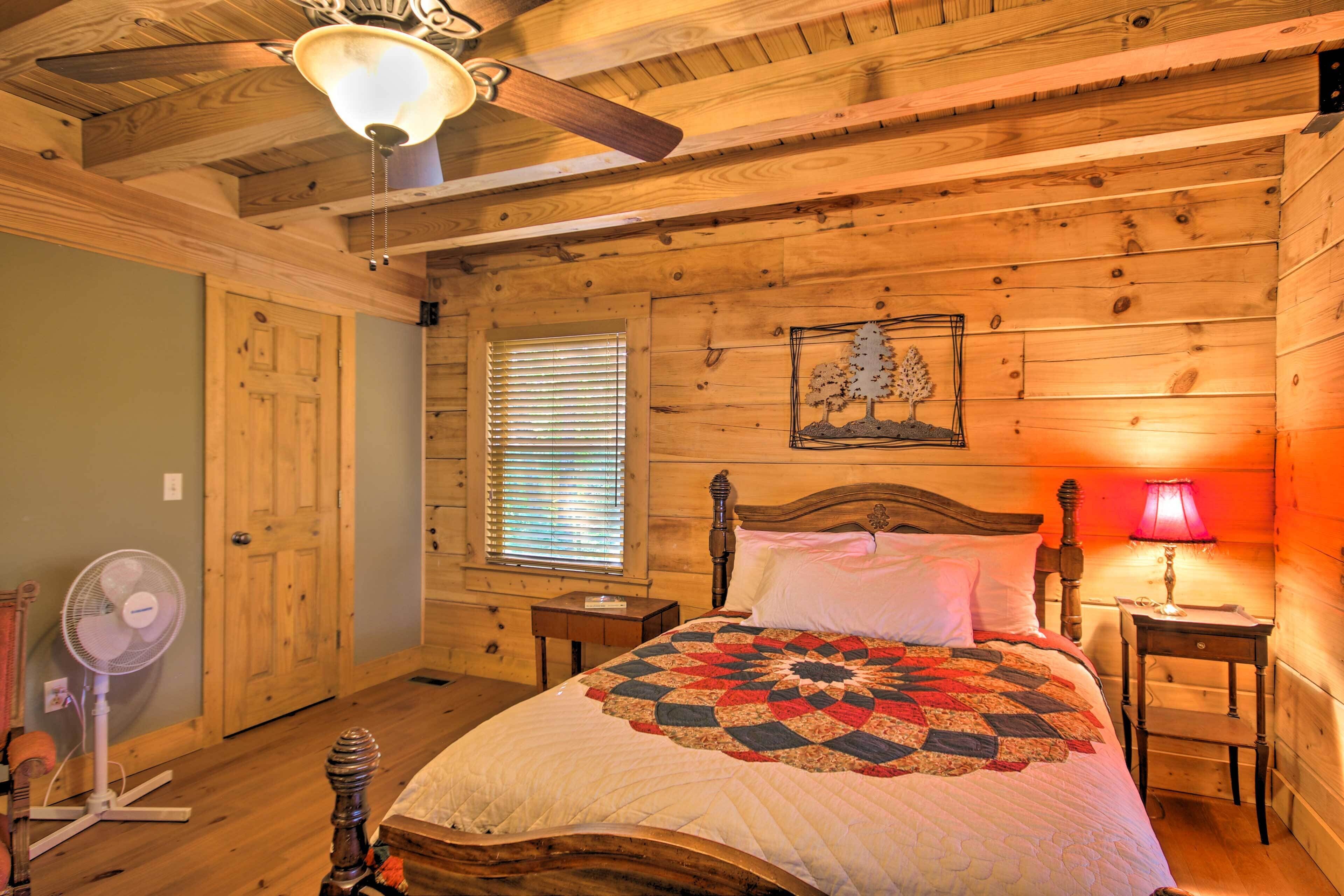 A wooden cabin bedroom with a bed, a ceiling fan, a window with blinds, and a nightstand with a lamp.