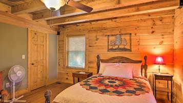 A wooden cabin bedroom with a bed, a ceiling fan, a window with blinds, and a nightstand with a lamp.