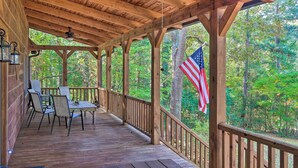 Eine überdachte Holzterrasse mit Tisch und Stühlen, umgeben von Bäumen und einer amerikanischen Flagge.