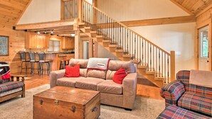 Ein geräumiger Wohnbereich mit einer Holztreppe, einem Sofa mit roten Kissen und einem Sessel mit Karomuster.