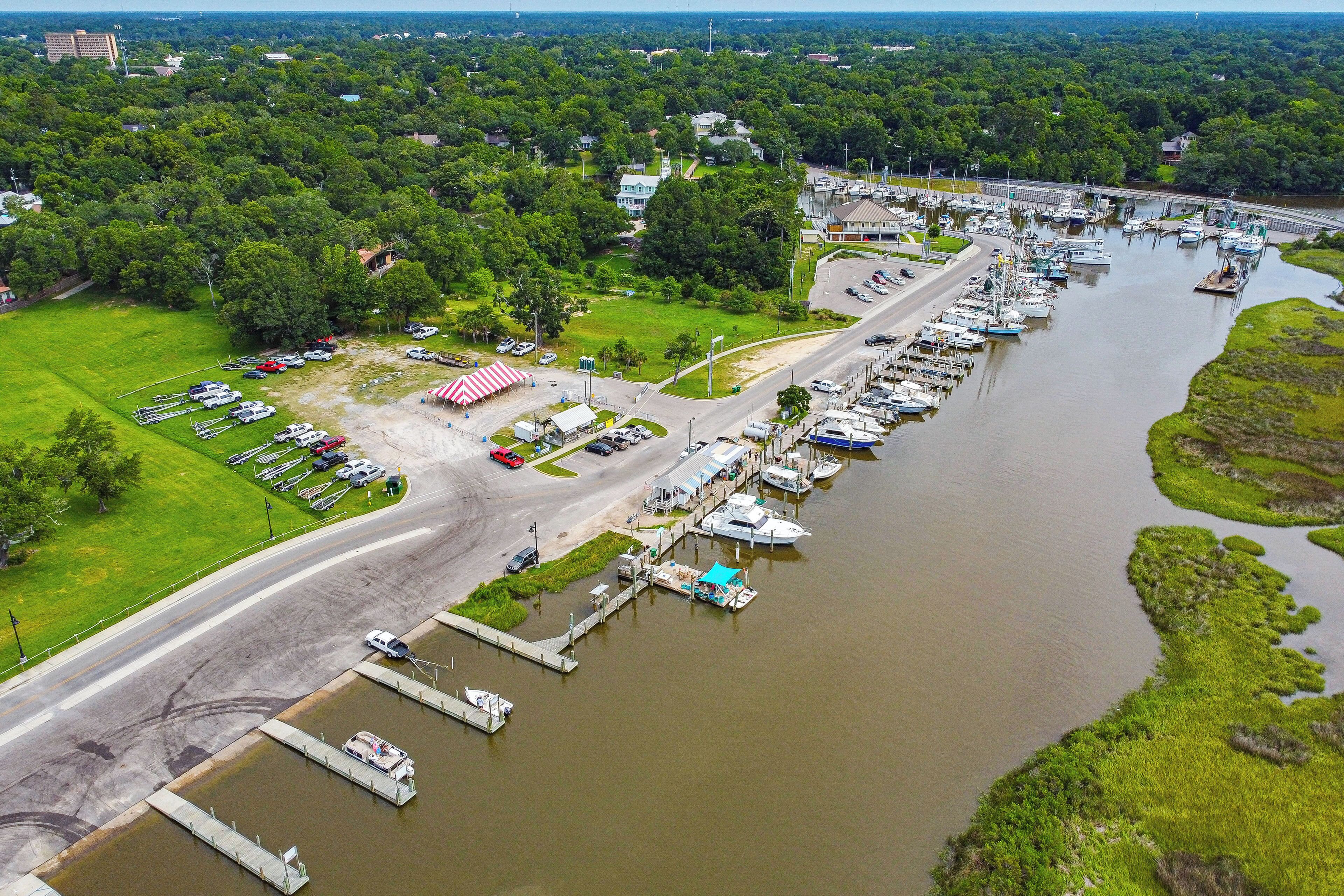 An aerial view of a marina with boats docked, a parking lot, and a grassy area with a small building.