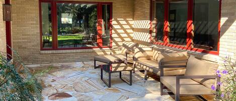 Une terrasse couverte avec un sol en pierre, du mobilier d’extérieur et de grandes fenêtres aux cadres rouges.