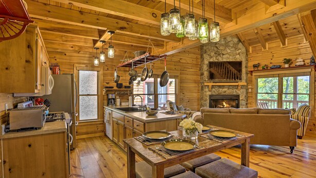 A rustic cabin interior with a wooden dining table set for four, a kitchen with a stone fireplace, and a cozy living area with a sofa and a chair. - Lovely Log Cabin + Bunkhouse w/ Views & Yard! (Cherry Log)