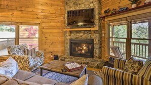 A cozy cabin interior with a stone fireplace, wooden walls, and a large window overlooking a deck. - Lovely Log Cabin + Bunkhouse w/ Views & Yard! (Cherry Log)