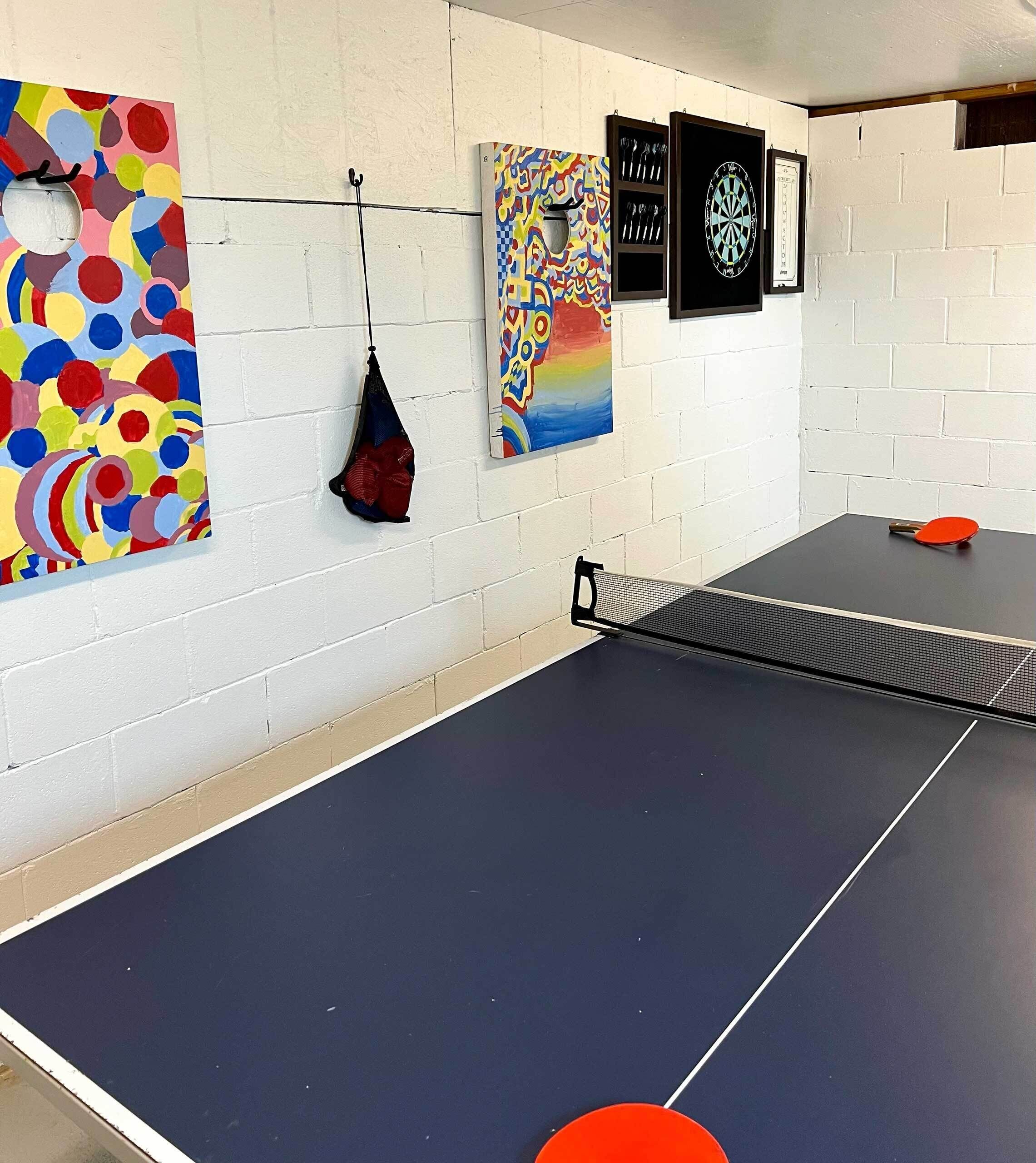 Une table de ping-pong avec un filet au centre, une peinture abstraite aux couleurs vives sur le côté gauche et un jeu de fléchettes sur le côté droit.