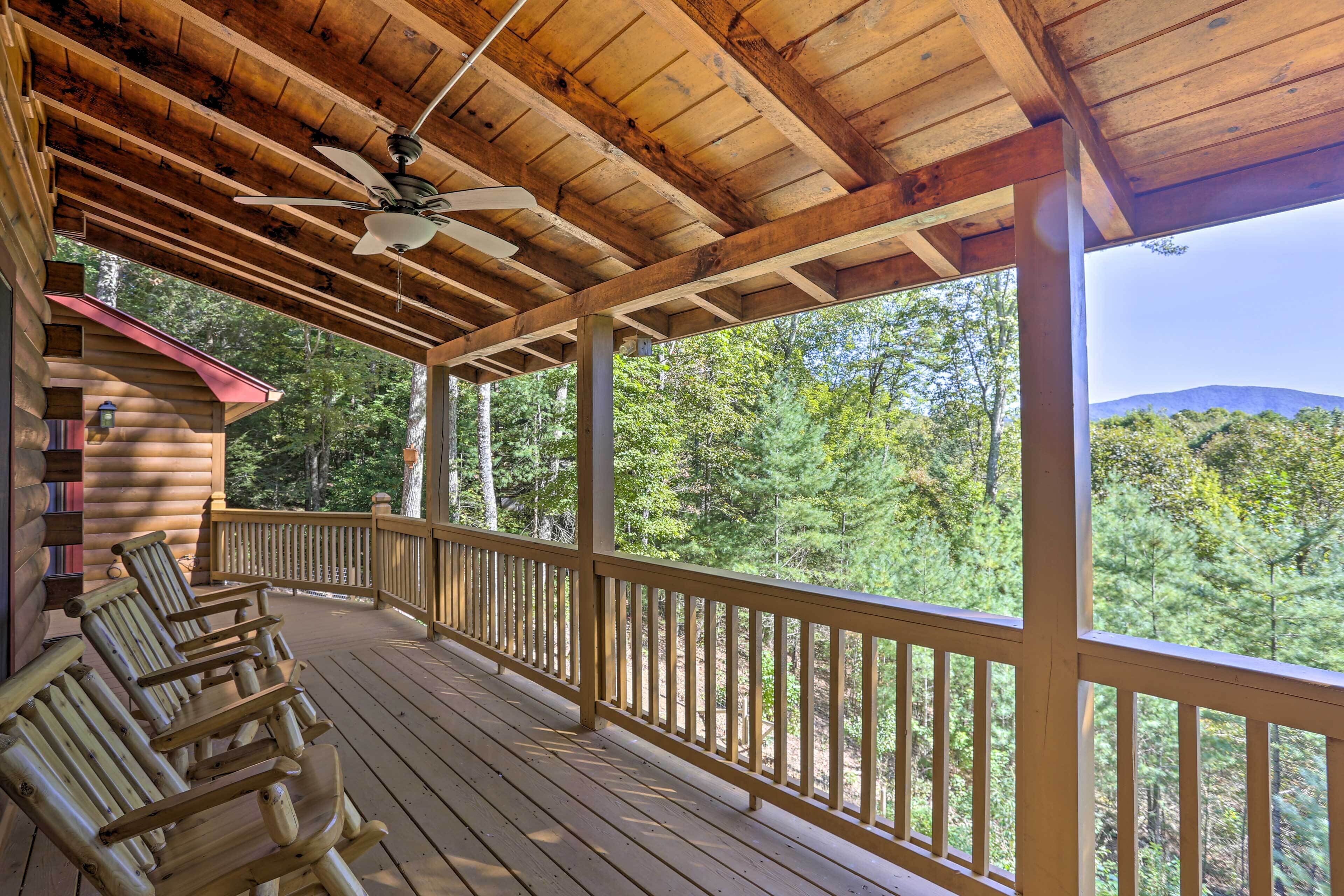 A wooden deck with rocking chairs, a ceiling fan, and a view of a forested area.