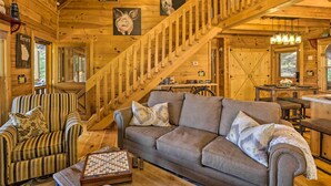 A cozy cabin interior with a wooden staircase, a striped armchair, a sofa, and a chessboard. - Lovely Log Cabin + Bunkhouse w/ Views & Yard! (Cherry Log)